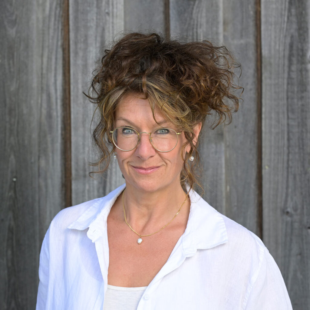 Frau mit lockigen braunen Haaren, Brille und weißer Bluse lächelt freundlich